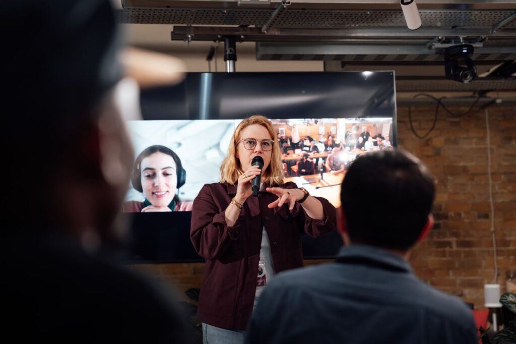 A public speaker on a mic addresses a crowd using multiple digital platforms for civic engagement, including two-way video conferences, a live audience, and feedback collection with CRM for the public sector